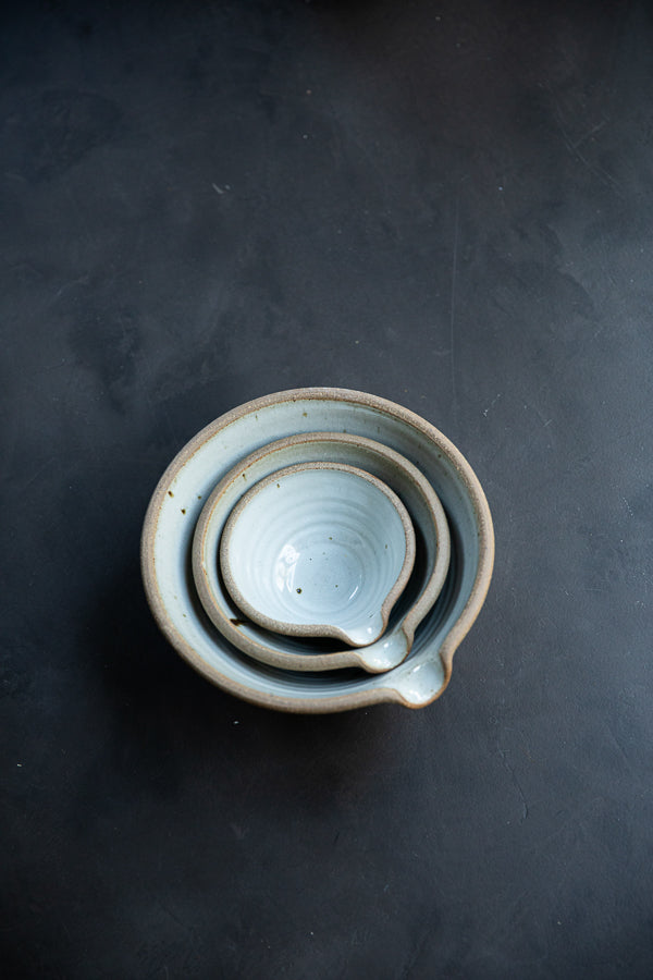 Handmade by the Leach Pottery Studio in St Ives. A set of three mixing bowls each finished with a lip for pouring. These bowls are glazed on the inside with a very pale duck egg blue dolomite glaze. The outside is unglazed for a natural and organic feel. Displayed on a dark background stacked inside each other