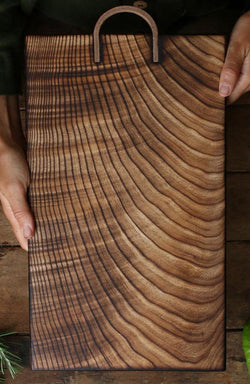 Handmade by Ambrose Vevers. These chopping boards are made from ash wood, which has been scorched with a naked flame and finished with food safe oil. A leather loop is at the top of the board for easy display and storage. The shape of this board is a rectangle.
